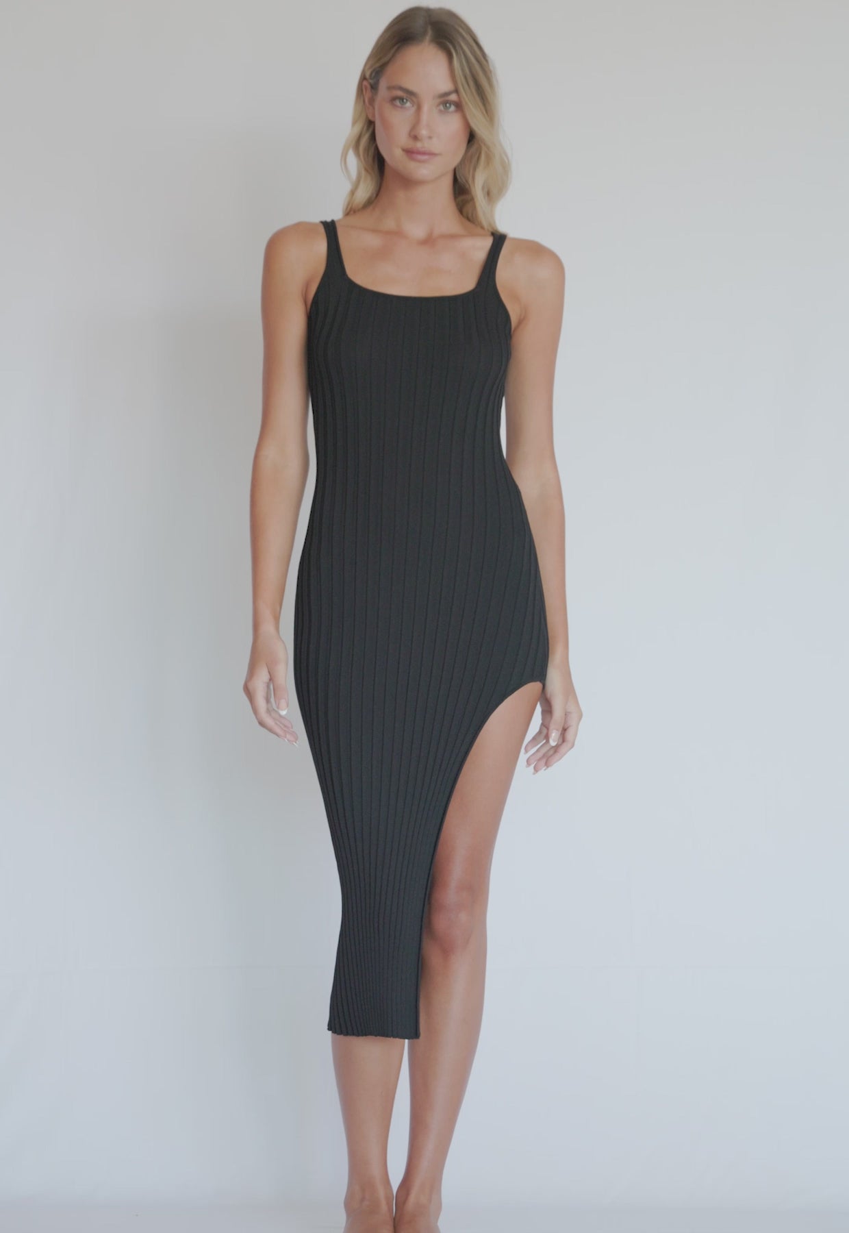 A blonde woman wearing a black dress with a side slit spinning in front of a white wall.