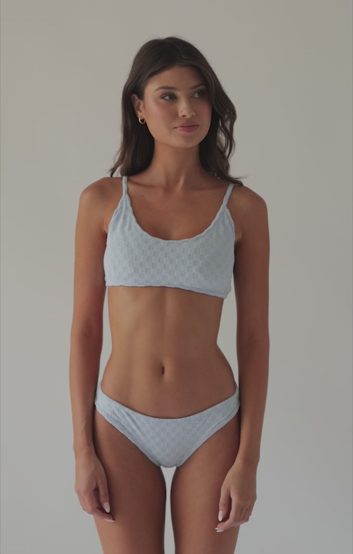 A brunette woman wearing a blue bikini spinning in front of a white wall.