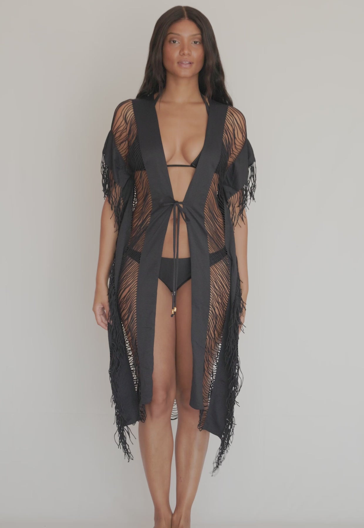 A woman wearing a black bikini and fringe cover up spinning in front of a white wall.