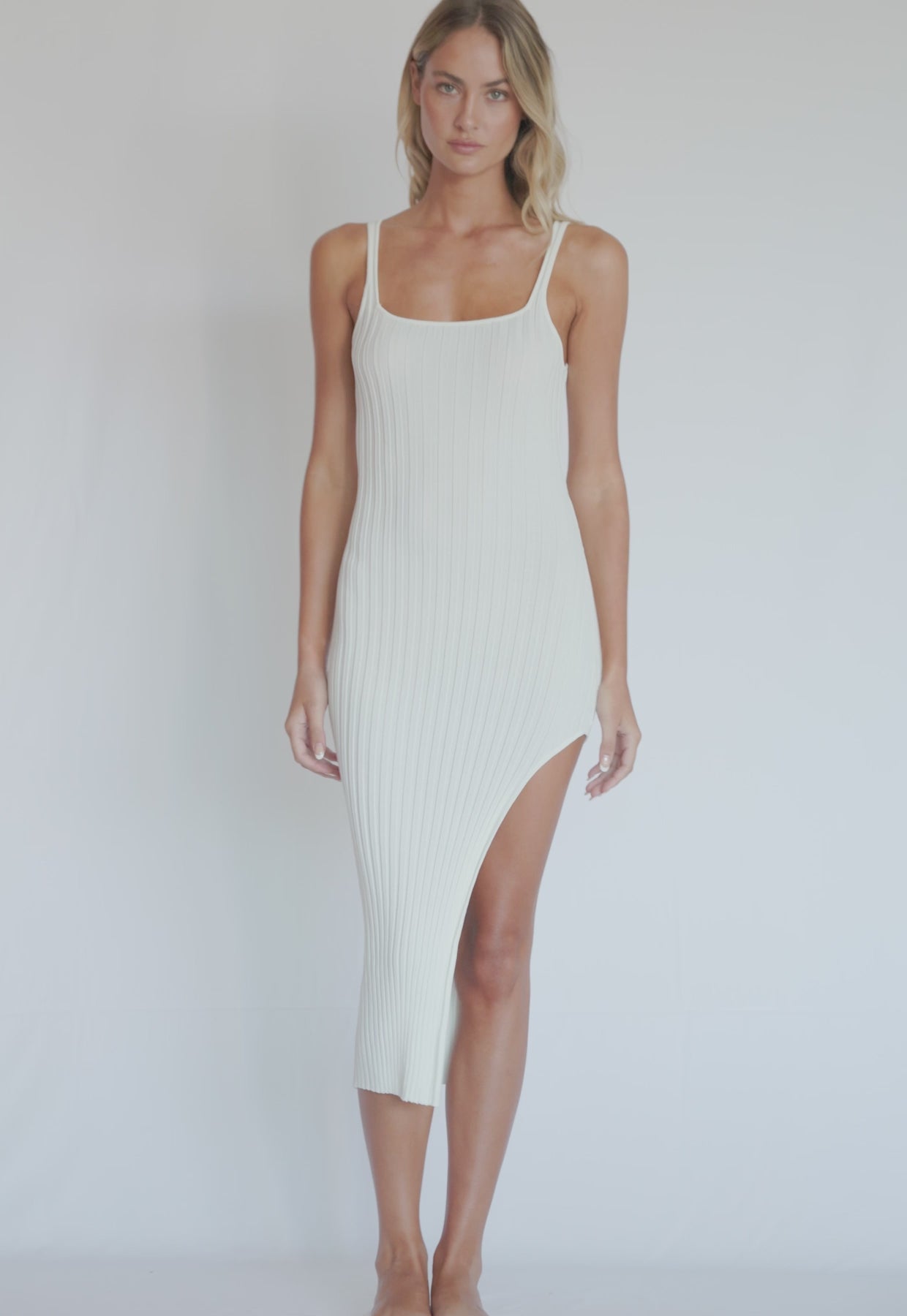 A blonde woman wearing a white dress with a side slit spinning in front of a white wall.