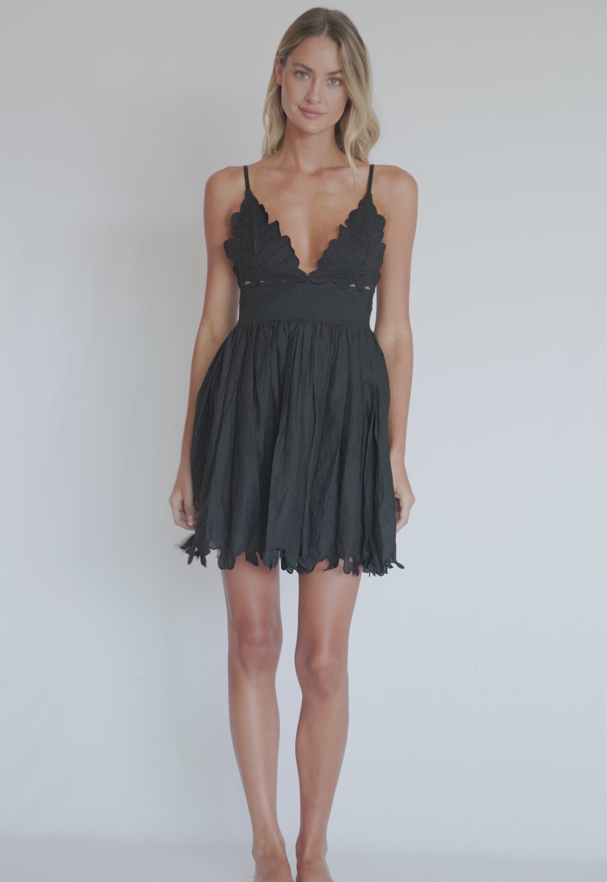 A blonde woman wearing a a short black dress spinning in front of a white wall.