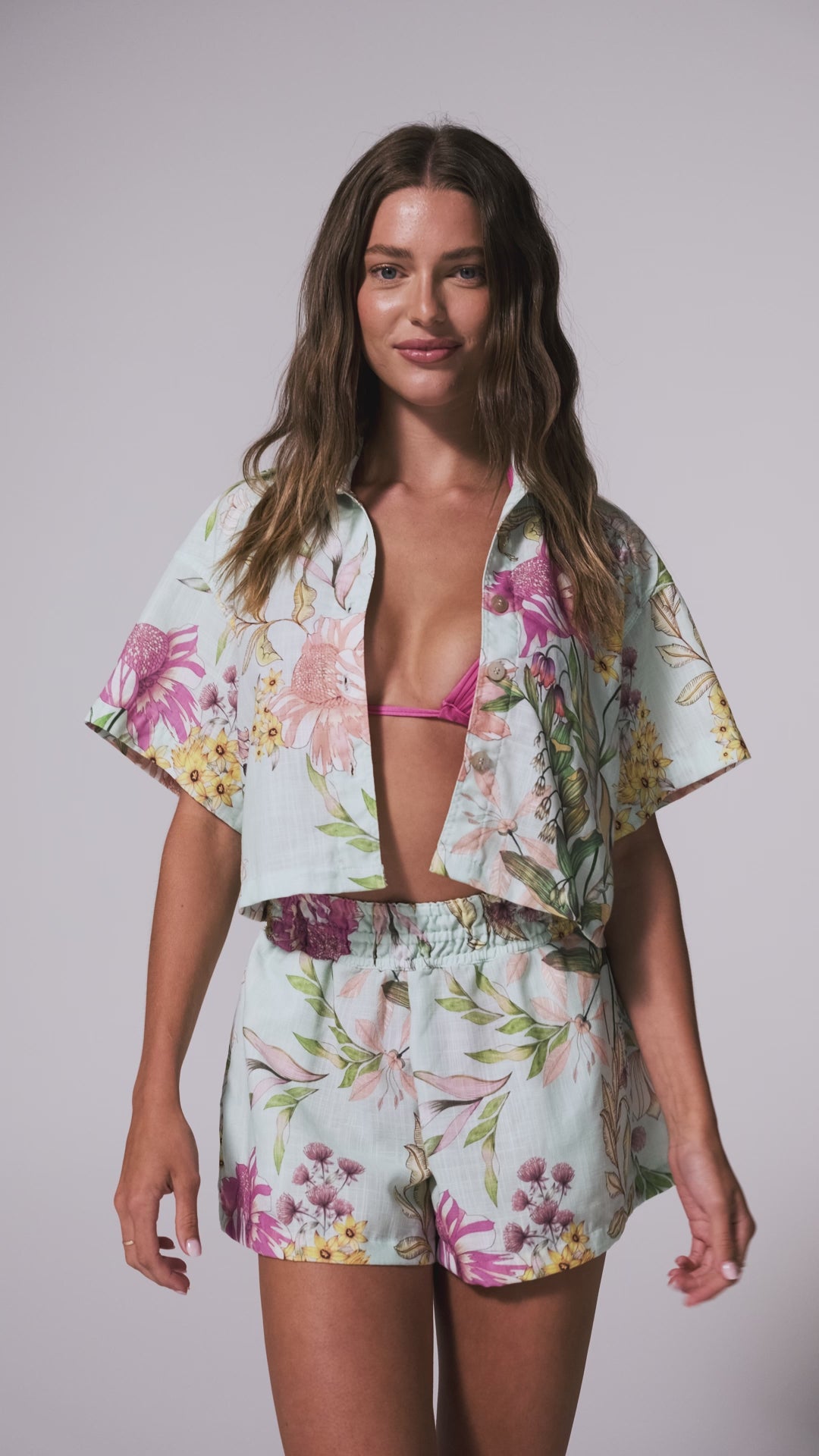 A woman wearing a pink bikini with a teal floral matching set of a short sleeve collared shirt and shorts spinning in front of a white background.