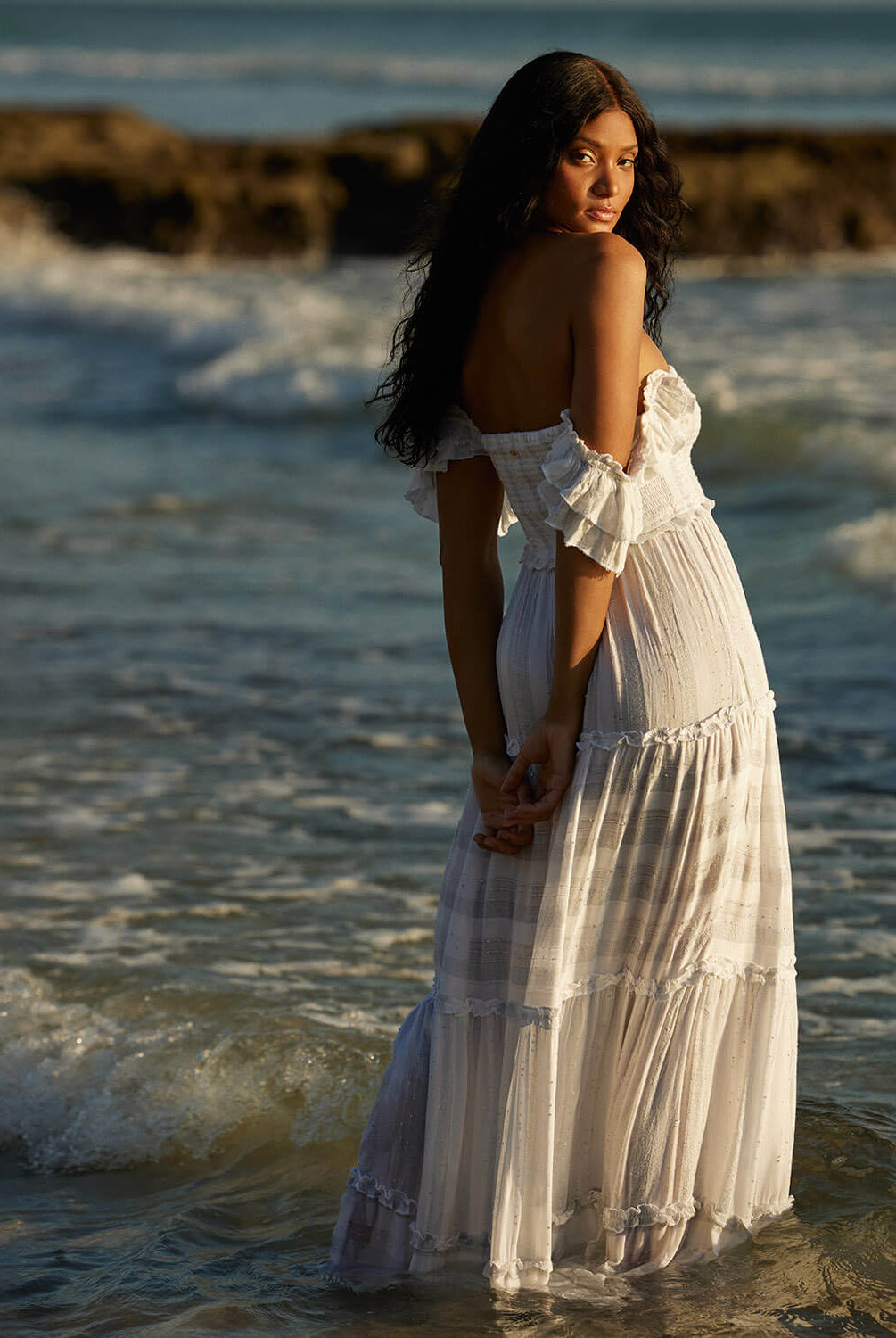 A woman wearing a white long dress standing on a beach in the ocean.