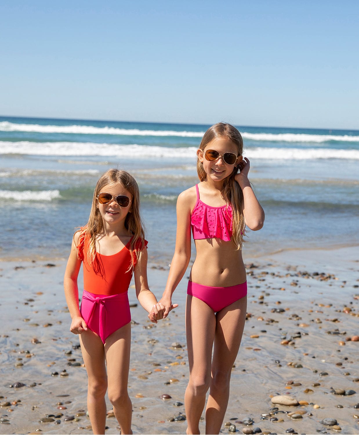 Two blonde girls wearing pink and red swimsuits standing on the beach near the ocean.
