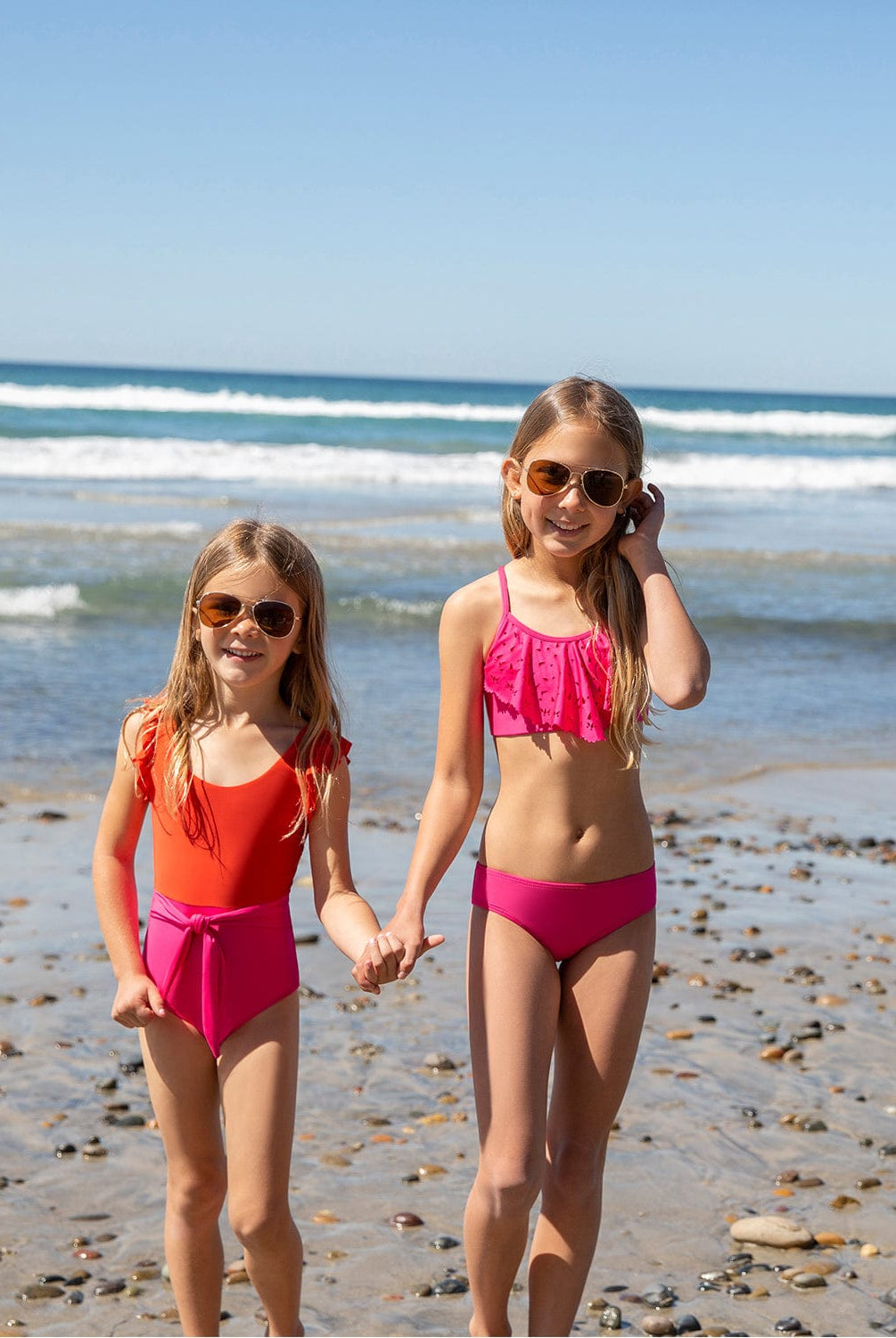 Two blonde girls wearing pink and red swimsuits standing on the beach near the ocean.