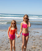 Two blonde girls wearing pink and red swimsuits standing on the beach near the ocean.