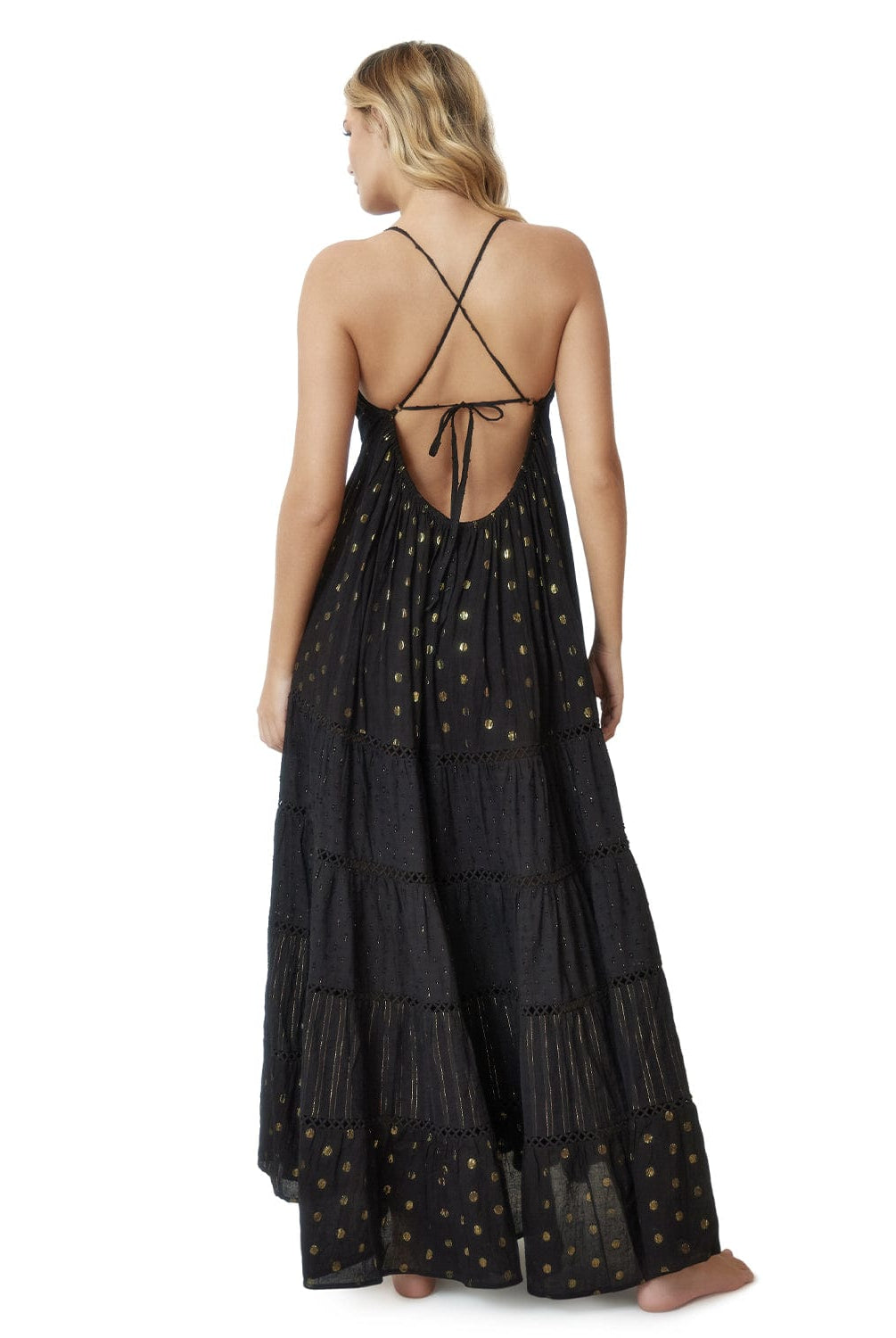Blonde woman wearing a black full length dress with shimmery gold accents and cross stitching embroidery facing backwards towards white wall.