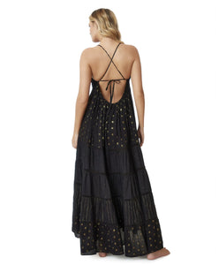 Blonde woman wearing a black full length dress with shimmery gold accents and cross stitching embroidery facing backwards towards white wall.
