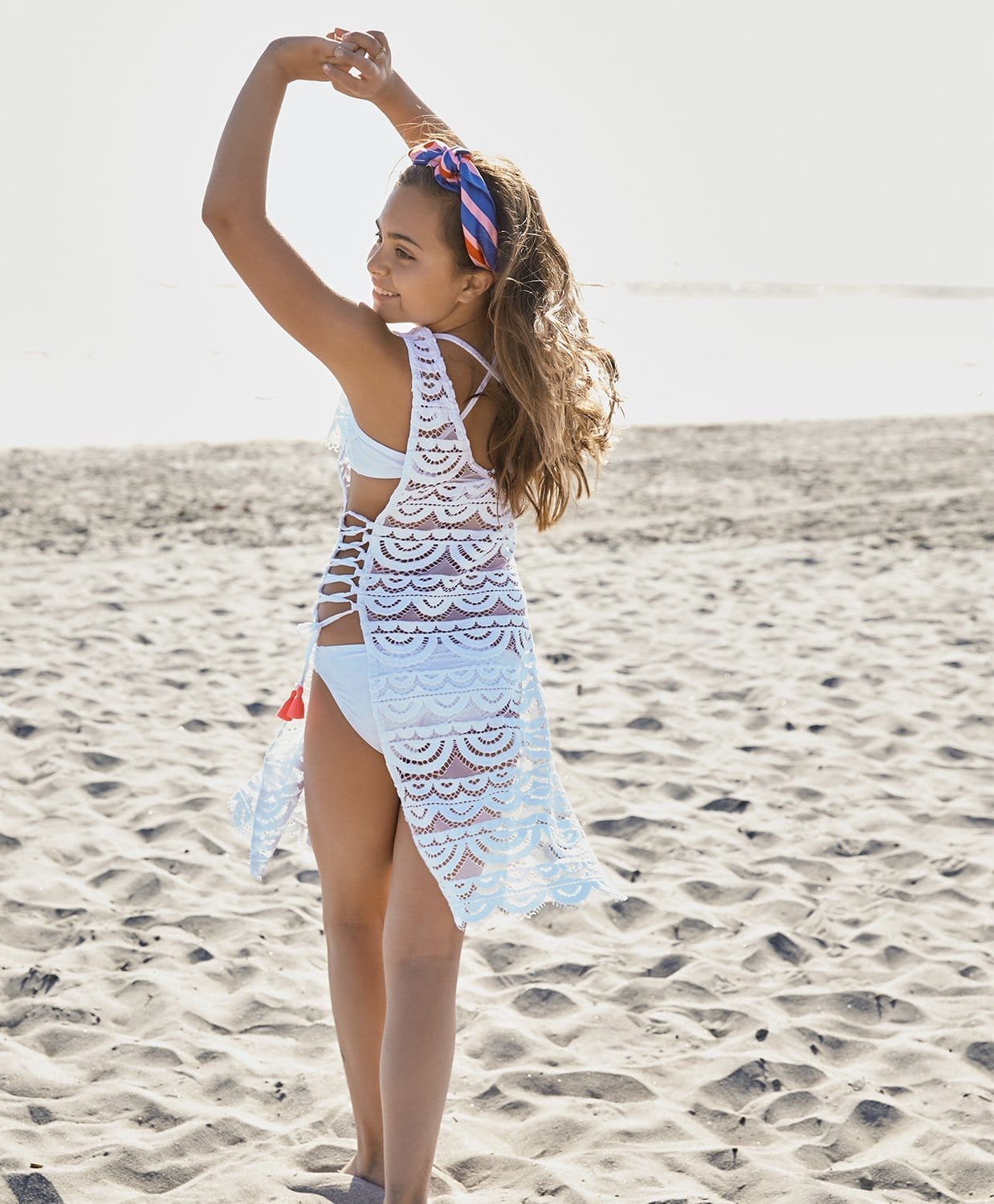 A brunette girl wearing a white lace cover up dress standing on a beach.