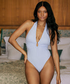 A woman wearing a light blue one piece swimsuit standing inside with a hand on her hip.