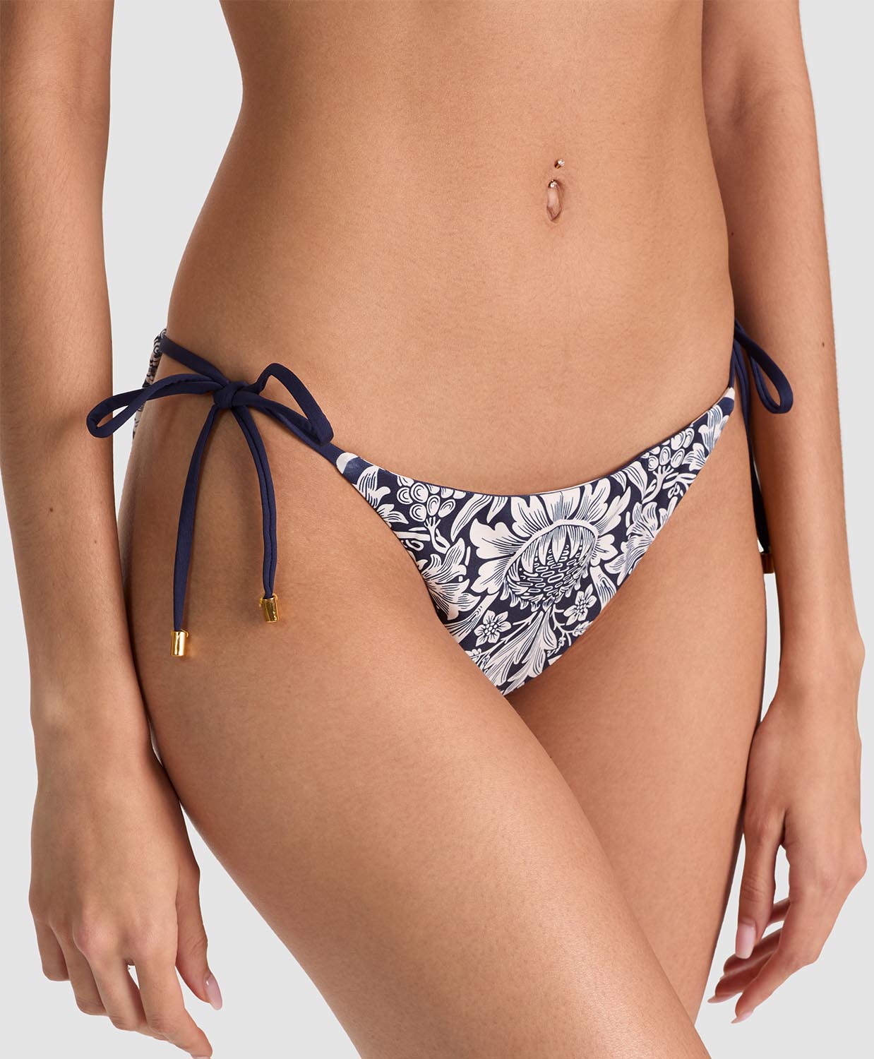 A close-up of a model wearing bikini bottoms with a navy blue and white floral pattern, featuring side-tie straps with gold-toned tips, against a light gray studio background.