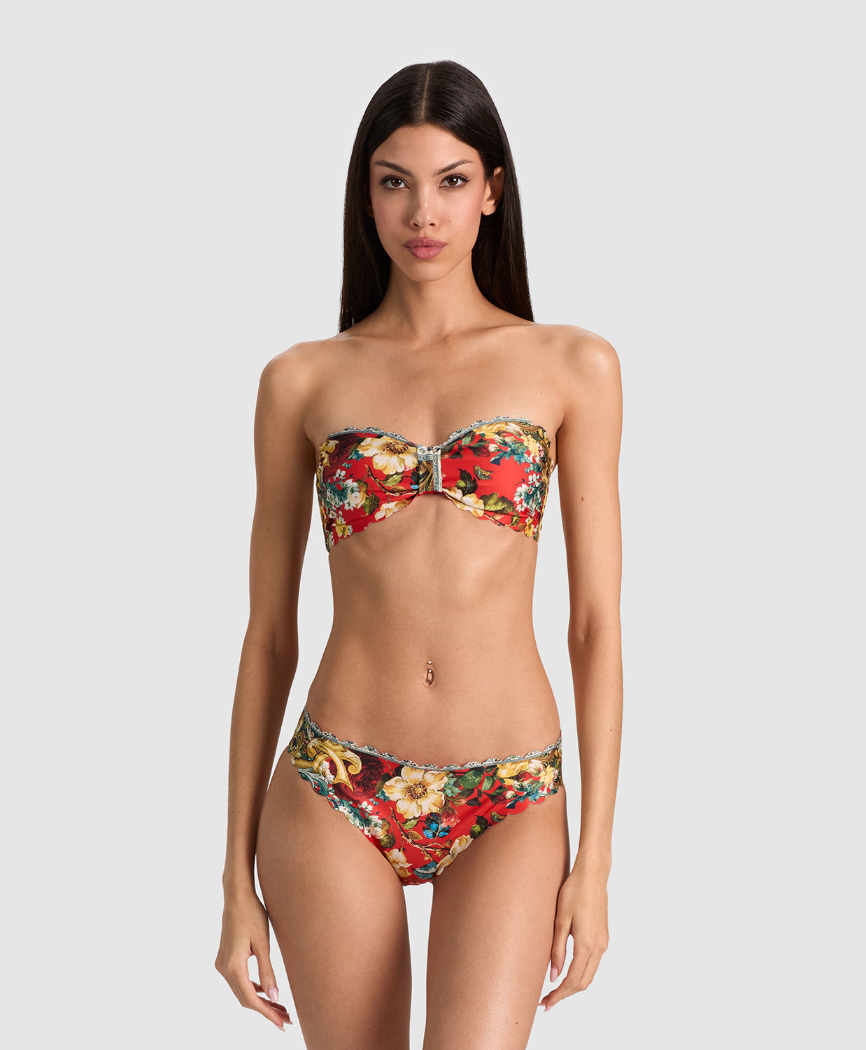 A model wearing a strapless bandeau bikini with a vibrant red, yellow, and green floral pattern, set against a solid light gray background.