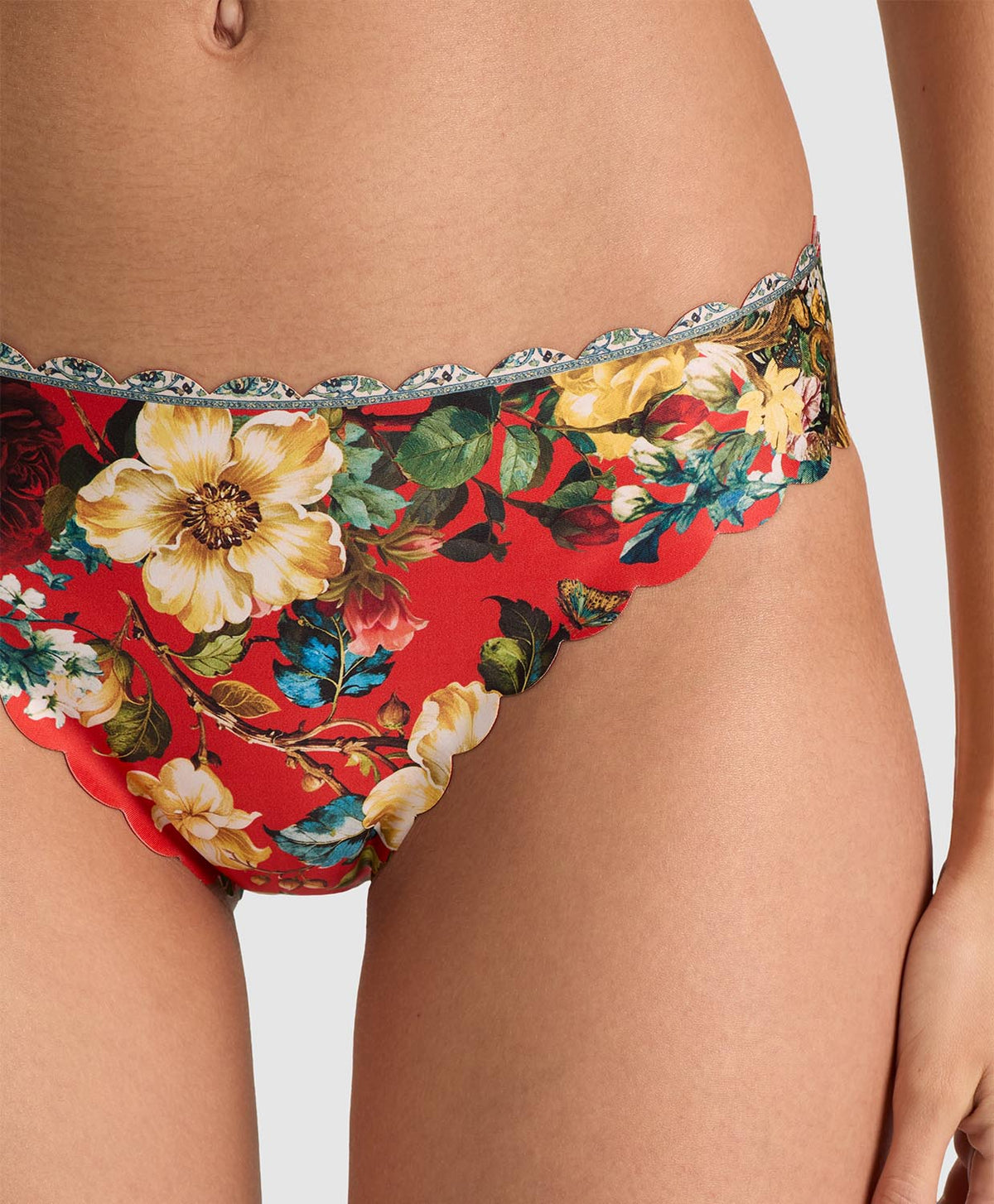 Close-up of a model wearing red floral bikini bottoms with a silver scalloped lace trim, featuring large yellow and white flowers with green leaves.
