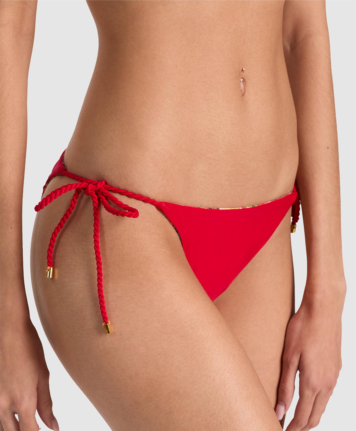 A close-up of a model wearing bright red bikini bottoms with side-tie rope details and small gold hardware accents, photographed against a light gray studio background.
