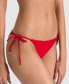 A close-up of a model wearing bright red bikini bottoms with side-tie rope details and small gold hardware accents, photographed against a light gray studio background.