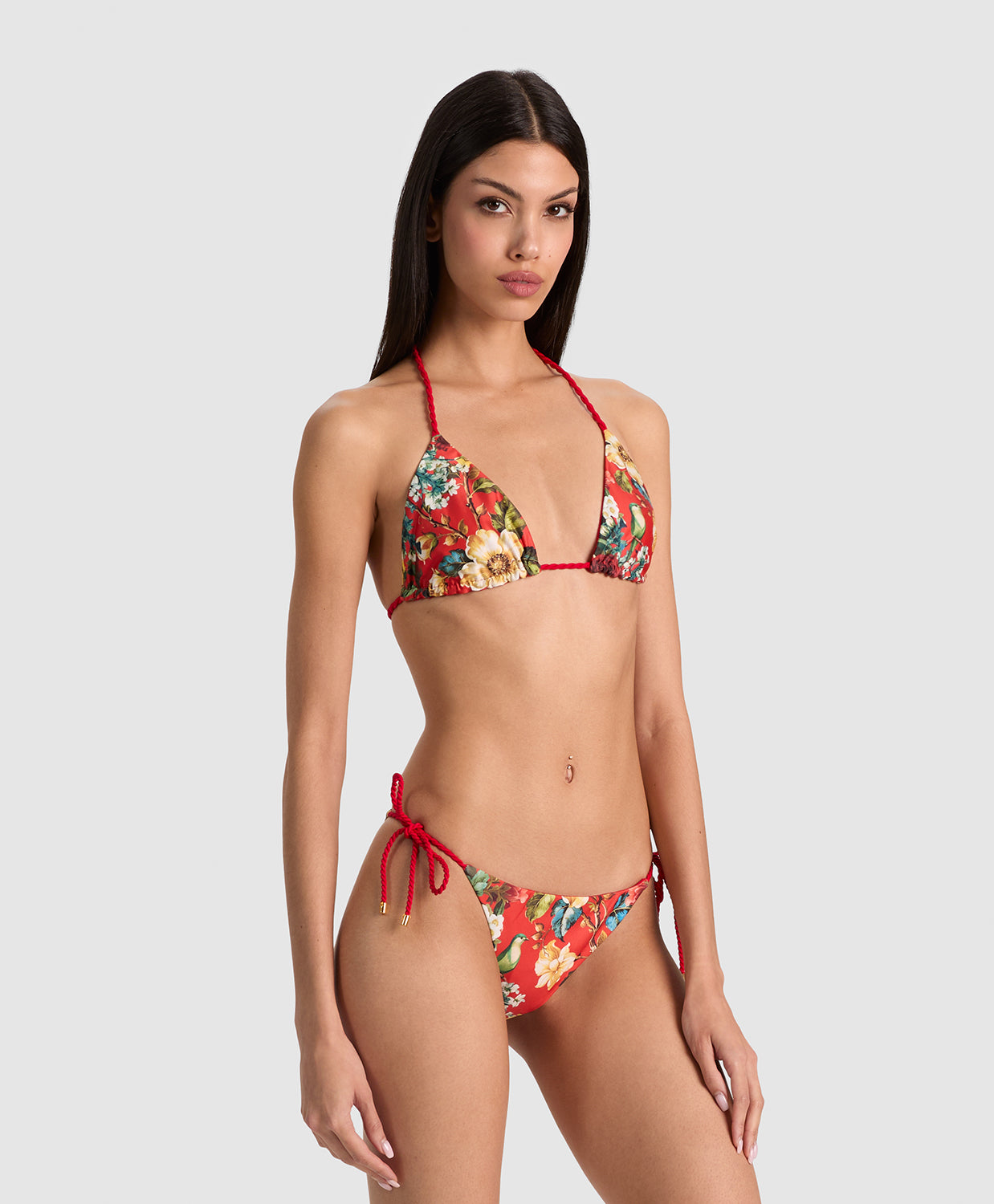 A woman modeling a two-piece bikini with a red string base and a colorful floral print, photographed against a solid light gray background.