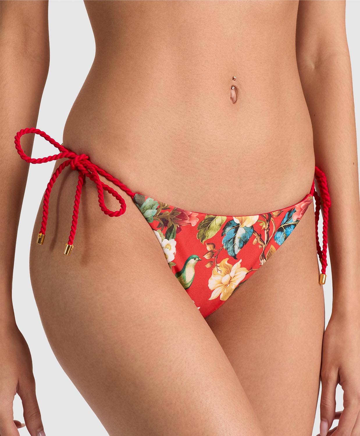A close-up of a model wearing red bikini bottoms with a tropical floral print and red rope side ties with gold-toned tips, shown against a light gray studio background.