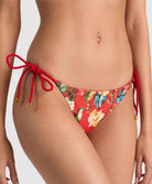A close-up of a model wearing red bikini bottoms with a tropical floral print and red rope side ties with gold-toned tips, shown against a light gray studio background.