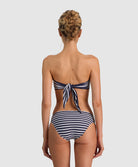 A woman viewed from the back wearing a navy and white striped bikini, featuring a tie-back top and high-waisted bottoms, set against a solid light gray background.
