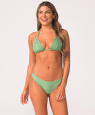 Woman wearing a green bikini with macrame detailing stands in front of white wall.