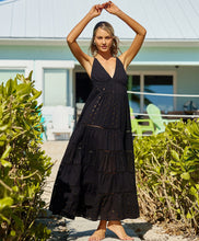 Blonde woman wearing a black full length dress with shimmery gold accents and cross stitching embroidery stands in front of beach house.