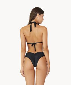 Brunette woman wearing a black halter bikini with net macramé details & shimmery gold accents facing backwards towards a white wall.