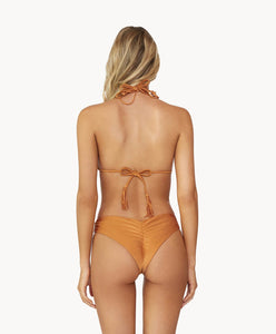 Blonde woman wearing an orange macrame triangle bikini facing backwards towards a white wall.