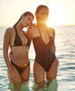 Brunette woman wearing a black halter bikini with net macramé details & shimmery gold accents and blonde woman wearing a black one piece swimsuit standing in the ocean.