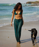 A woman wearing a green bikini top and matching pants walking on the beach with a dog.