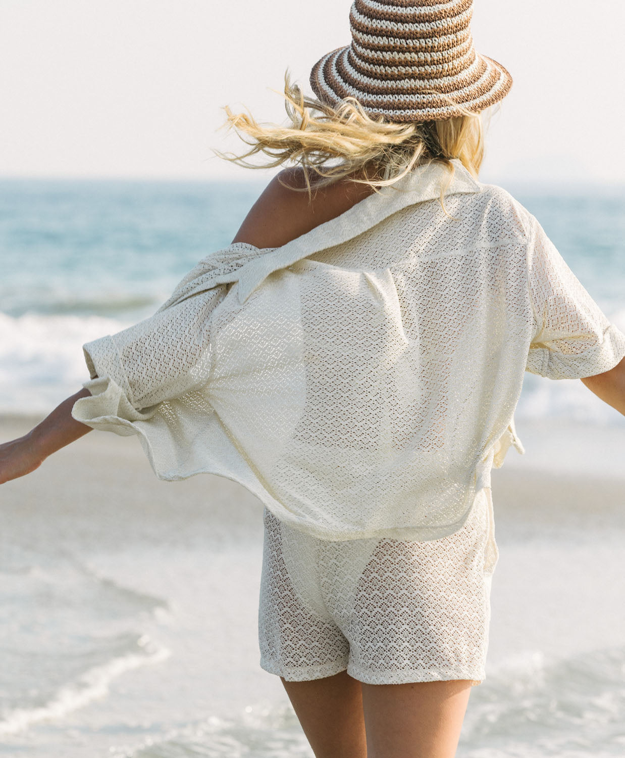Woman in a beige outfit and straw hat standing on a beach with ocean view