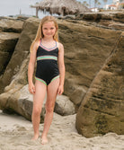 Young girl in a black swimsuit with colorful trim standing on a rocky beach.