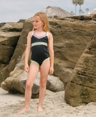 Young girl in a black swimsuit with colorful patterns standing on a rocky beach.