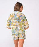 A woman wearing a matching floral shirt and shorts facing towards a white wall.