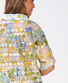 A woman wearing a floral crochet shirt facing towards a white wall.