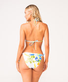 A woman wearing a triangle top bikini with a floral print facing towards a white wall.