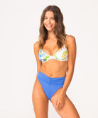 A woman wearing a white floral bikini top and blue high waist bottoms standing against a white wall.