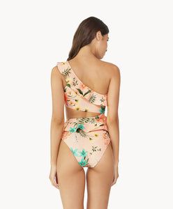 A brunette woman wearing a one piece in a tropical print facing towards a white wall