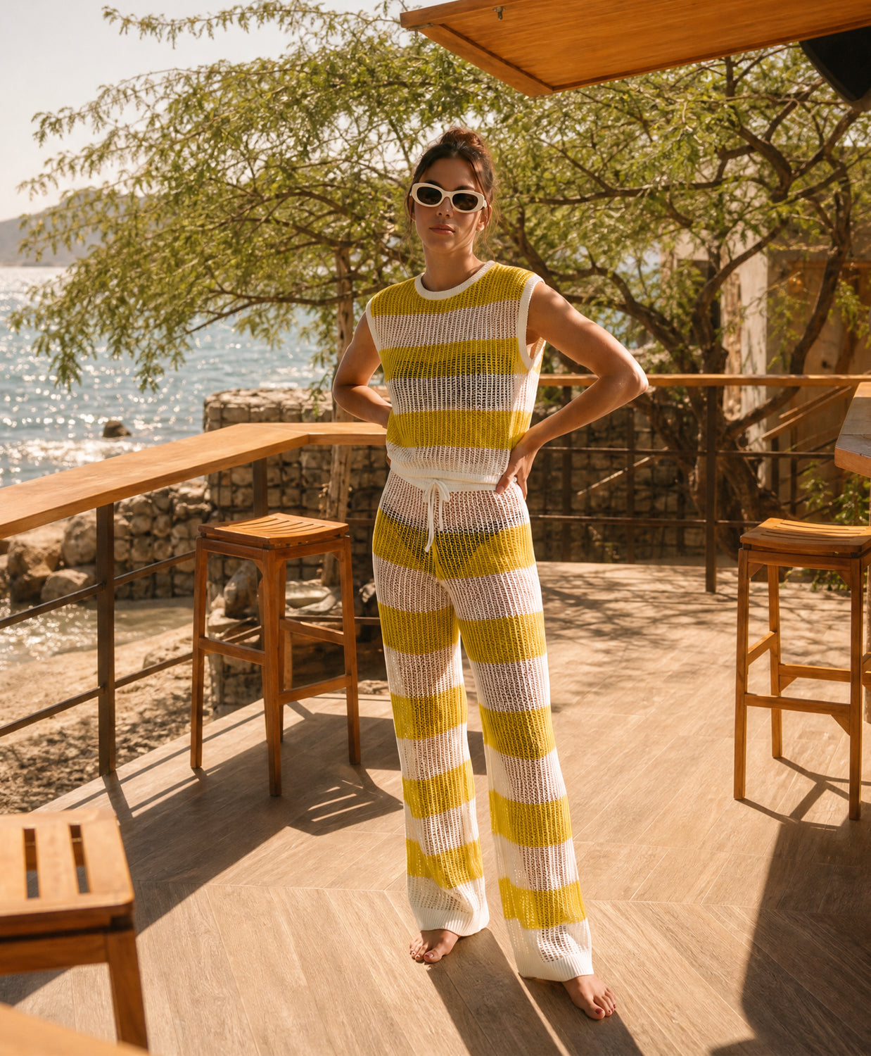 A woman stands barefoot on a wooden deck by the water, wearing a sleeveless ribbed knit top and matching wide-leg pants in alternating yellow and white horizontal stripes.