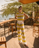 A woman stands barefoot on a wooden deck by the water, wearing a sleeveless ribbed knit top and matching wide-leg pants in alternating yellow and white horizontal stripes.