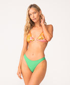 A woman wearing an orange triangle bikini top and green bikini bottom standing against a white wall. 