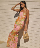 A woman wearing a long, colorful, strapless dress leaning against a brick wall.