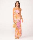 A woman wearing a long, strapless, pink and orange dress with a floral print standing against a white wall.