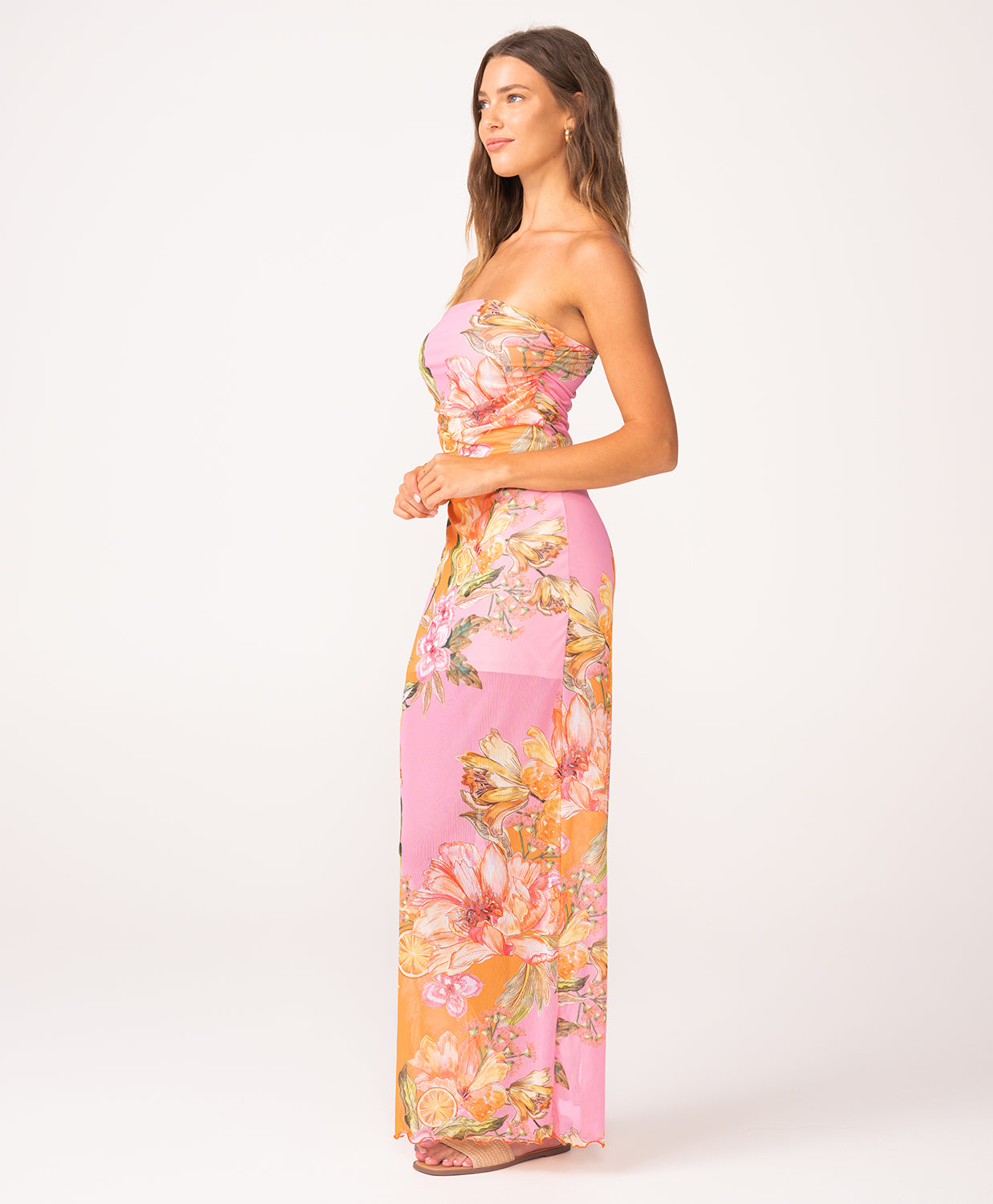 A woman wearing a long, strapless, pink and orange dress with a floral print standing against a white wall.