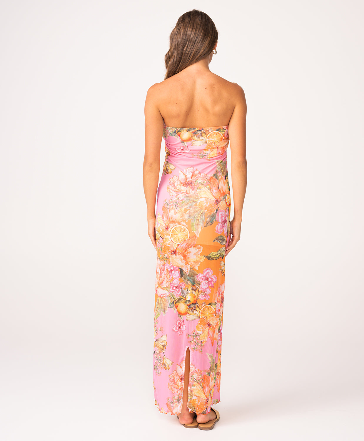 A woman wearing a long, strapless, pink and orange dress with a floral print facing towards a white wall.