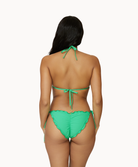 A woman wearing a green bikini facing towards a white wall. 