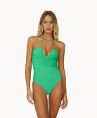 A blonde woman wearing a green one piece swimsuit standing in front of a white wall. 