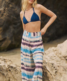 A woman wearing a navy triangle bikini top and tie dye pants standing outside near a rock wall.