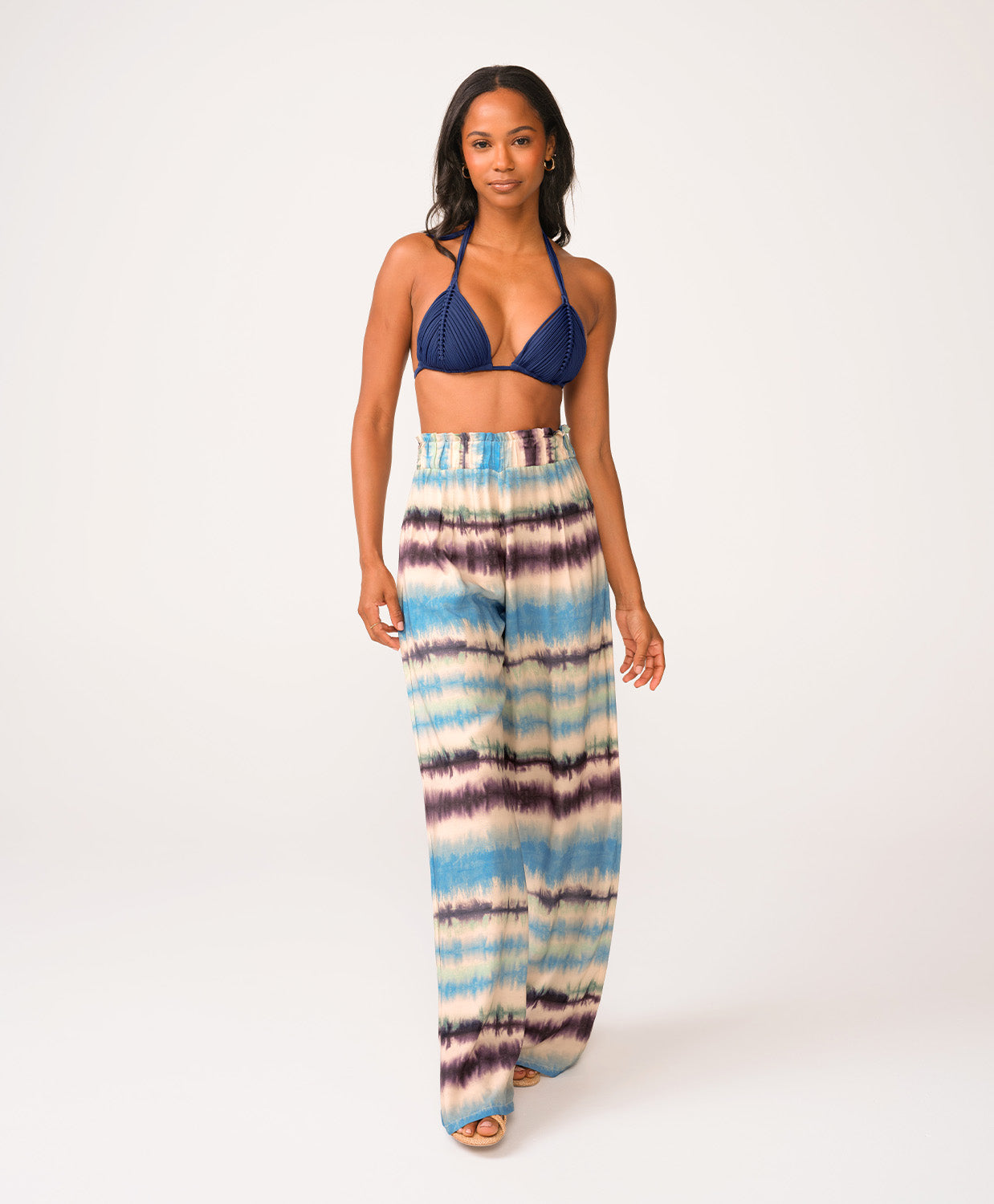 A woman wearing a navy triangle bikini top and blue tie dye pants standing against a white wall.