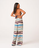 A woman wearing a navy triangle bikini top and blue tie dye pants standing against a white wall.