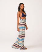 A woman wearing a navy triangle bikini top and blue tie dye pants standing against a white wall.